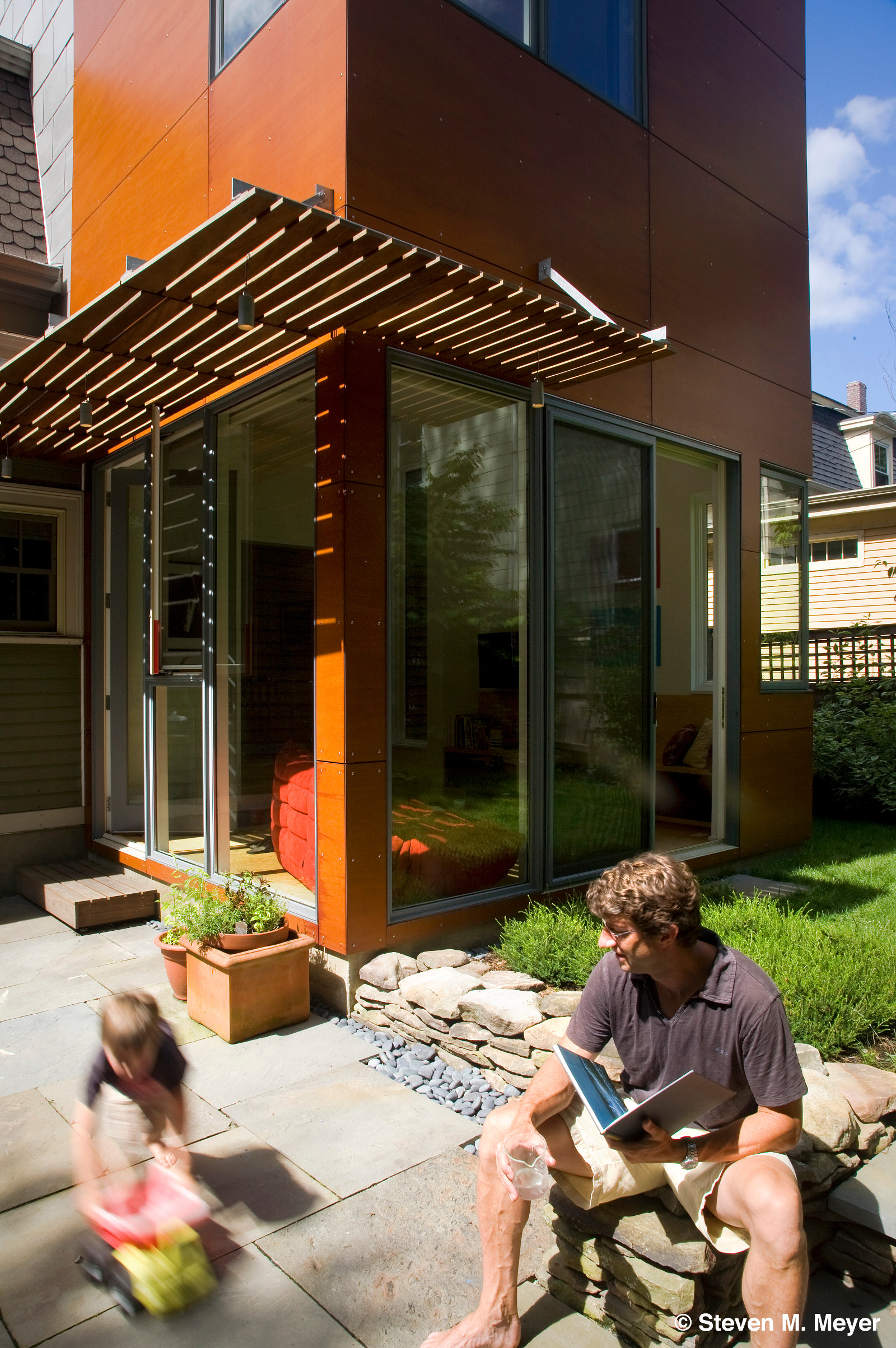 Modern residential addition with wood slat trellis, wood-veneer composite panel siding, large sliding doors, and floor-to-ceiling windows.