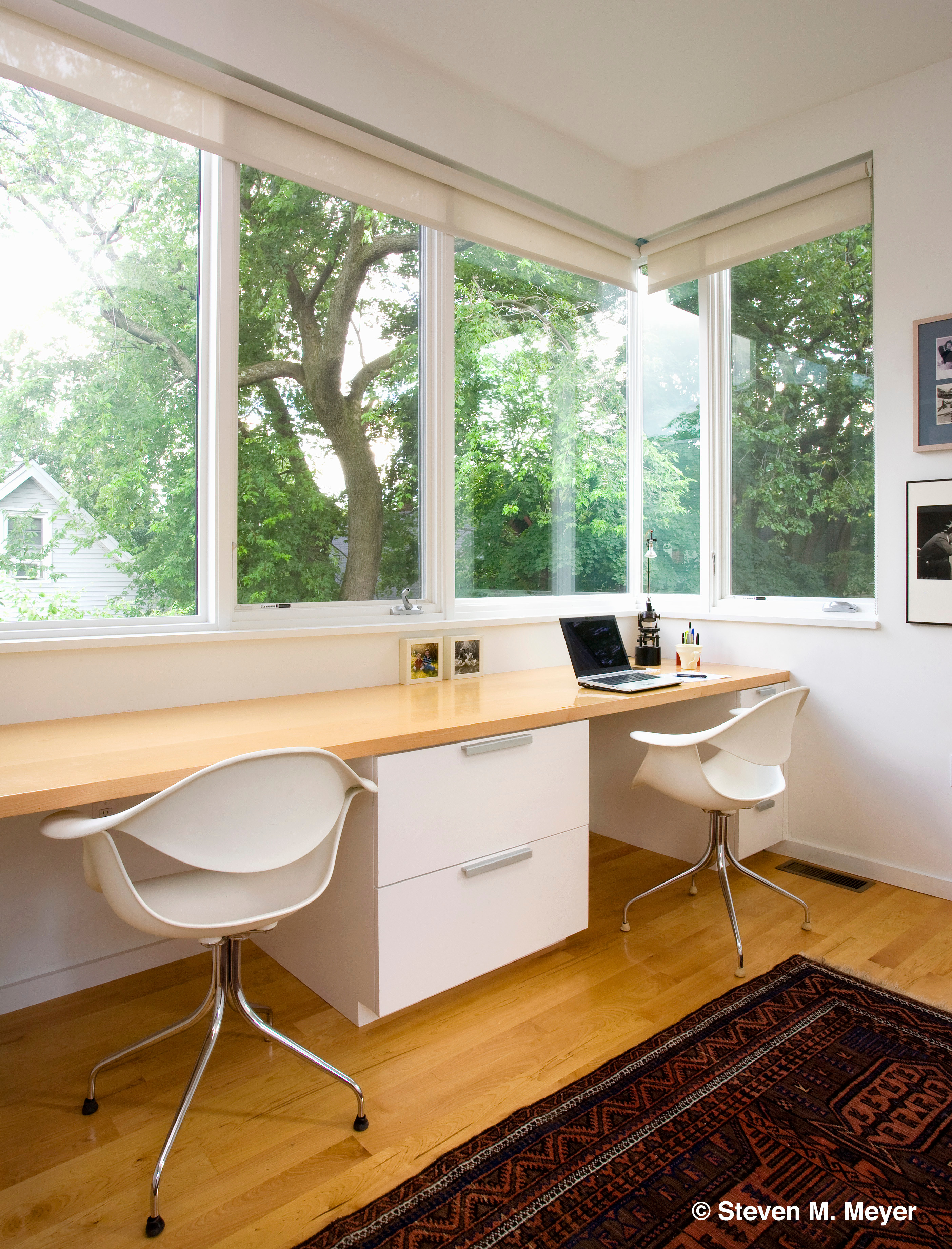 Home office with large corner window, maple desk, white cabinets.