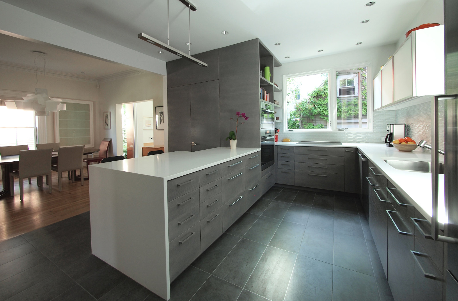 Modern kitchen with windows, gray cabinets, white Caesarstone counters, translucent glass upper cabinets, glass backsplash, and dark gray porcelain tile floor.