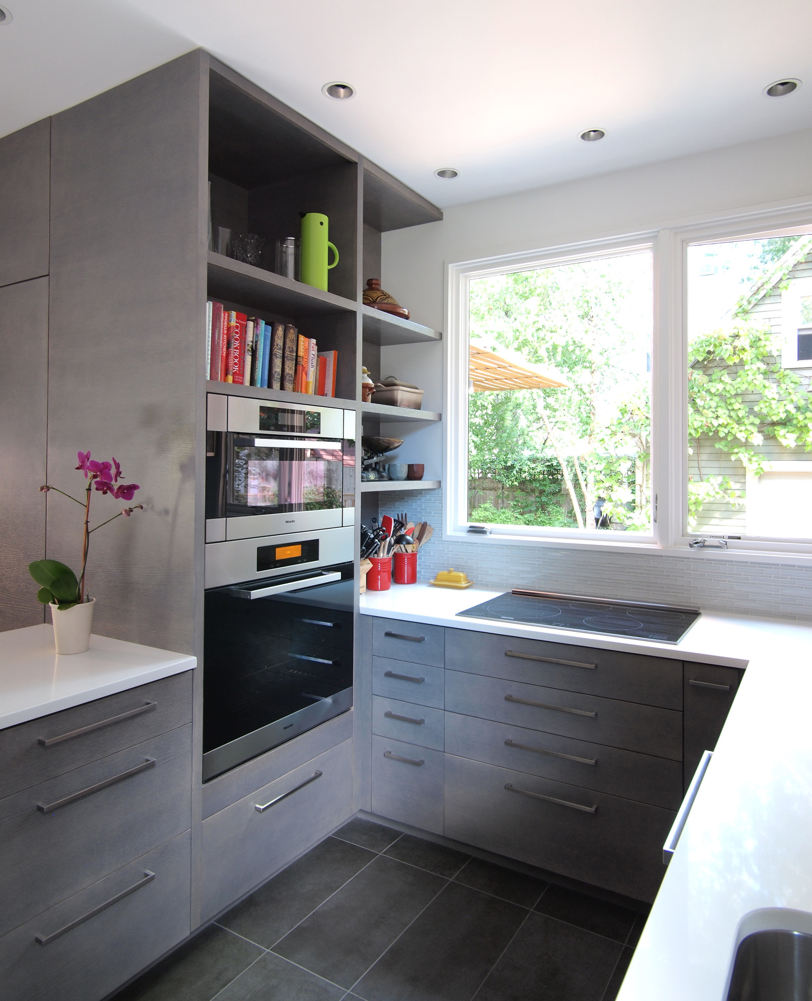 Modern custom kitchen with wall oven, built-in microwave, open shelving, induction cooktop, gray cabinets, white counter, and large window over cooktop.